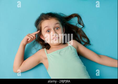 Schelmische Kindermädchen in stylischer Kleidung, Grimaka, macht Gesichter, Gesten, legt ihren Zeigefinger auf ihren Tempel, schaut in die Kamera. Reise, Reise, k Stockfoto