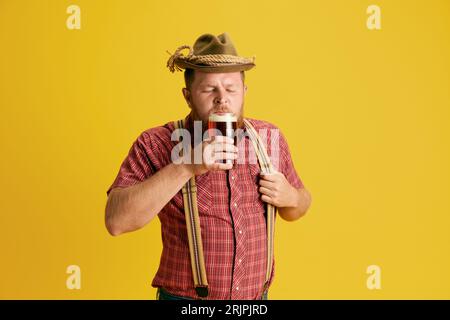 Bärtiger Mann in kariertem Hemd und Hut, der köstlich mit Bier vor gelbem Studiohintergrund schlürfen kann. Bayerische Traditionen Stockfoto