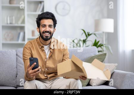 Porträt eines glücklichen Käufers in einem Online-Shop, der Mann erhielt das Paket pünktlich und qualitativ hochwertige Lieferung hispanic lächelt und schaut auf die Kamera, hält das Telefon in seinen Händen, schreibt eine positive Bewertung. Stockfoto