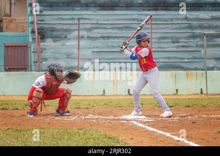 Ein kleiner Junge in einer Baseballuniform steht bereit, um bei einem Baseballspiel einen Ball zu schlagen, während ein Catcher und ein Schiedsrichter hinter dem Backstop zusehen Stockfoto