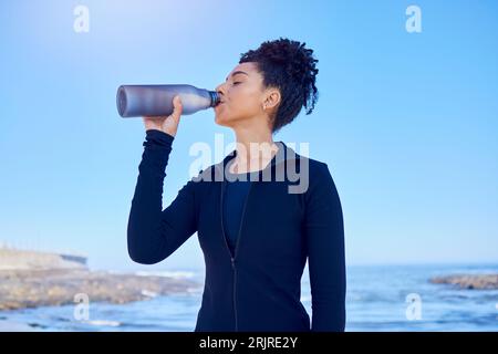 Fitness, Frau und Trinkwasser am Strand zum Laufen, Gesundheit oder Morgenkardio am blauen Himmel am Meer. Bewegung, Hydration und durstige Läuferin Stockfoto