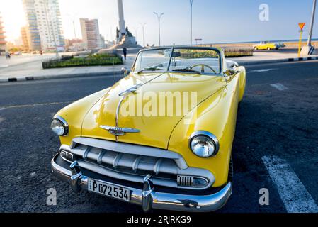 Ein gelber, alter Chevy aus den 1950er Jahren parkte an einer Straße in Malecon, Vedado, Kuba. Auf dem Hintergrund ein anderes gelbes Cabriolet vorbeifahrend Stockfoto