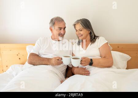 Smiling old caucasian husband and wife lies in bed, cheers with cups of coffee, enjoy good morning Stockfoto