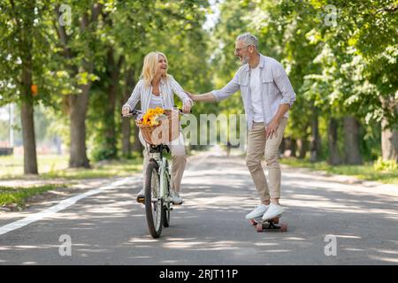 Active Mature Couple Riding Bike And Skateboard On Path In Park Stock Photo
