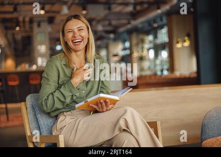 Lächelnde Managerin, die während der Zusammenarbeit auf einem Stuhl sitzt und sich während des Arbeitstages Notizen macht Stockfoto