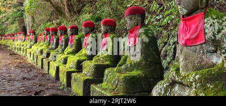 Moss bedeckte Jizo-Statuen, die rote Hüte und Lätzchen trugen, im Kanmangafuchi-Abgrund in Nikko, Japan Stockfoto