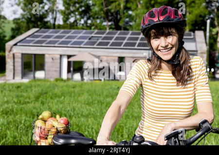 Porträt einer glücklichen Frau mit Fahrrad und Bio-Obst auf einer Wiese vor einem Haus Stockfoto