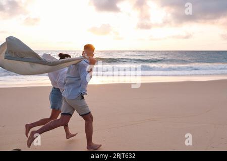 Mann, der mit einer Frau läuft, die am Strand eine Decke hält Stockfoto