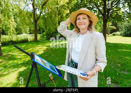 Lächelnde reife Frau, die im Park einen Hut mit Aquarellfarben trägt Stockfoto