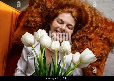 Junge Rothaarige Frau, die einen Haufen Tulpenblüten zu Hause hält Stockfoto