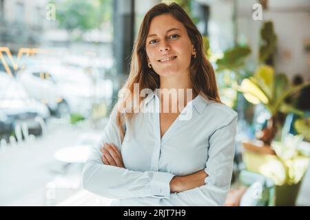 Glückliche, selbstbewusste junge Frau, die im Coffee Shop mit gekreuzten Armen steht Stockfoto