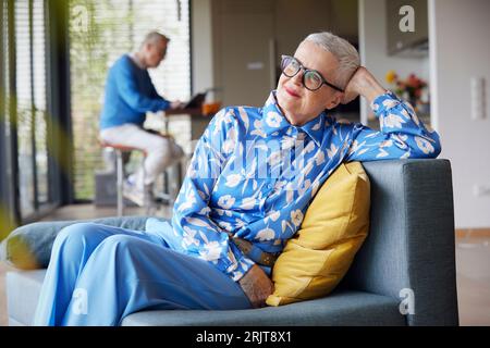 Entspannte ältere Frau, die zu Hause auf dem Sofa sitzt, mit Mann im Hintergrund Stockfoto