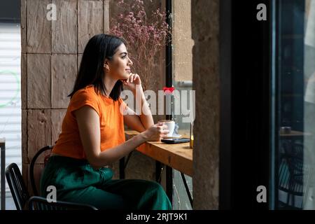 Nachdenkliche junge Frau, die mit einer Tasse Kaffee aus dem Fenster im Café schaut Stockfoto