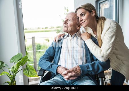 Lächelnde blonde Frau, die sich um den Vater kümmert, der zu Hause im Rollstuhl sitzt Stockfoto