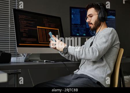 Junger Computerprogrammierer, der am Schreibtisch ein Smartphone benutzt Stockfoto