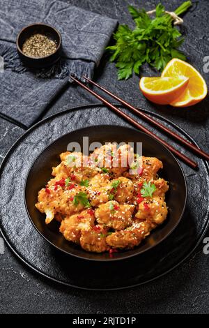 Knusprige orangefarbene Blumenkohlröschen, bestreut mit Sesamsamen, Chiliflocken und Petersilie in schwarzer Schüssel auf Betontisch, vertikale Ansicht Stockfoto