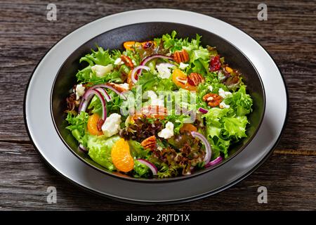 Mandarinen-Orangen-Salat mit Salatmischung, getrockneten Preiselbeeren, Pekannüssen, roter Zwiebel und Fetakäse in schwarzer Schüssel auf dunklem Holztisch Stockfoto