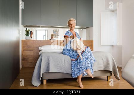 Glückliche reife Frau mit Hut sitzt auf dem Bett in der Wohnung Stockfoto