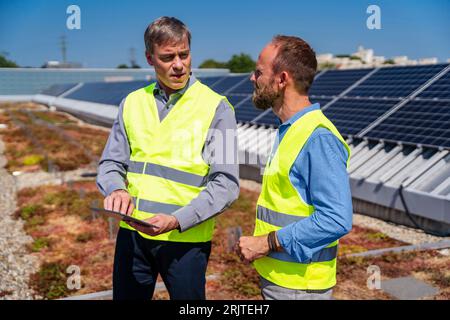 Zwei Männer mit Tablet-PC, die reflektierende Westen tragen, sprechen auf dem Dach eines Firmengebäudes mit Solarpaneelen Stockfoto