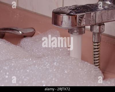 Rosafarbene Badewanne bis zum Rand gefüllt: Metallischer Wasserhahn fließt mit Wasser und reichlich Schaum Stockfoto