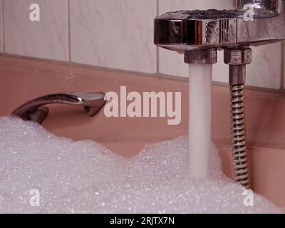 Rosafarbene Badewanne bis zum Rand gefüllt: Metallischer Wasserhahn fließt mit Wasser und reichlich Schaum Stockfoto