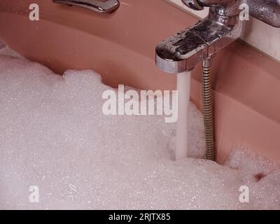 Rosafarbene Badewanne bis zum Rand gefüllt: Metallischer Wasserhahn fließt mit Wasser und reichlich Schaum Stockfoto