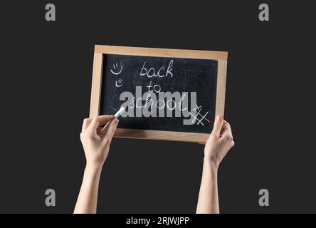 Weibliche Hände halten Kreide und Tafel mit Text ZURÜCK ZUR SCHULE auf schwarzem Hintergrund Stockfoto