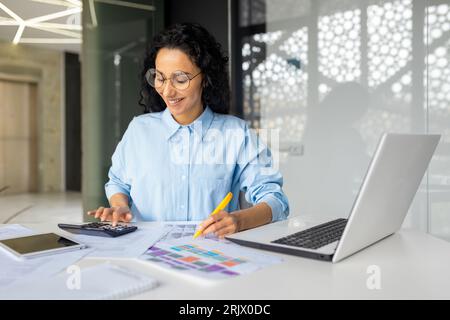 Erfolgreiche Geschäftsfrau Finanzierin auf Papier arbeitet mit Verträgen, berichtet über Arbeiten am Arbeitsplatz, hispanic Smiling verwendet Taschenrechner und Laptop, weibliche Mitarbeiterin zufrieden mit Leistungsergebnissen Stockfoto