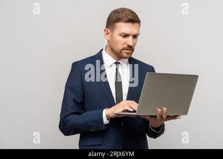 Ein seriöser Geschäftsmann im Anzug, der auf einen Notebook-Bildschirm auf grauem Hintergrund schaut. Stockfoto