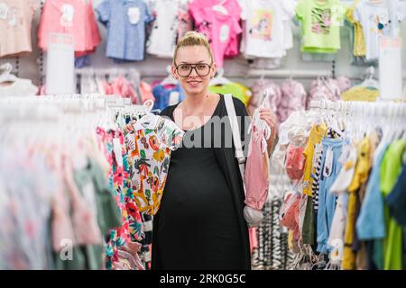 Eine schwangere Frau entscheidet, Kinderkleidung im Store. Stockfoto