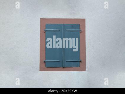 Braunes, traditionelles Fenster mit zwei blauen geschlossenen Holzjalousien auf leerem weißem Steinhintergrund. Stockfoto