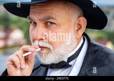 Nahaufnahme des seriösen, stilvollen Hipster-Großvaters im Ledermantel, der draußen raucht. Älterer Großvater, Opa-Rentner, Rentner-Konzept. Stockfoto
