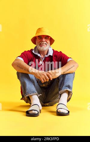 Portrait of attractive cool man sitting on floor wearing sunglasses and hat isolated over bright yellow color background. Stockfoto