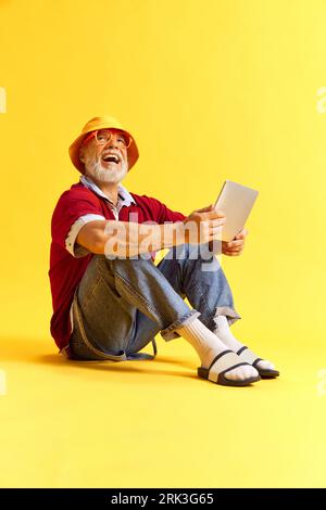 Ganzer Blick auf die Körpergröße eines attraktiven, fröhlichen und lustigen Mannes, der auf dem Boden mit Tablet sitzt und Spaß auf einem rosa pastellfarbenen Hintergrund hat Stockfoto