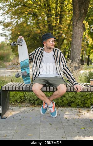 Portrait of stylish adult male who sitting at extreme park near skateboard, summer vibe. resting Stockfoto