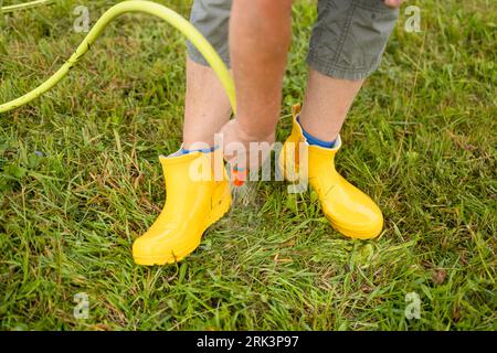 Nahaufnahme der Hände einer Frau wäscht gelbe Gummistiefel nach der Arbeit im Boden mit einem Gartenschlauch aus dem Schlamm Stockfoto