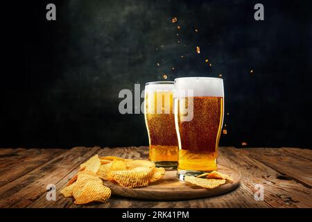 Zwei Gläser schäumendes, kühles Lagerbier mit Pommes Frites Vorspeisen auf Holztisch vor dunklem Hintergrund Stockfoto