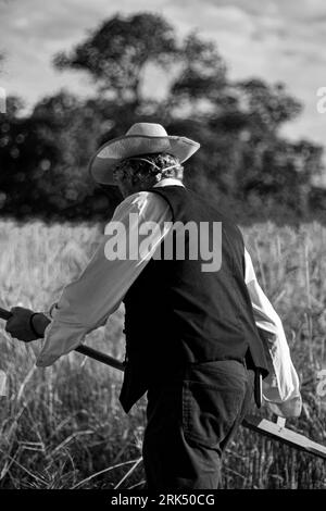 Ein schwarz-weißes Bild eines Reifen Mannes, der auf einem grasbewachsenen Feld steht und eine Sense hält Stockfoto