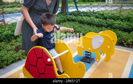 A cute two year old baby boy has fun playing on a colorful swing in the park, while his mom watches over his safety. Stock Photo