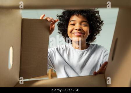Ansicht von der Innenseite des Kartons. Afrikanisches Mädchen, das die Lieferung auspackt und in die Schachtel schaut. Frau öffnet Karton. Eine Frau bekommt ein Paket, das sich die gelieferten Waren ansieht. Zufriedenes positives Feedback des Kunden Stockfoto