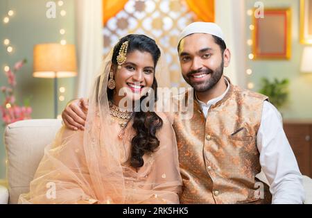 Happy smiling indain Muslim couple embracing by looking at camera while sitting on sofa at home - concept of traditional culture, Ramadan greeting and Stockfoto