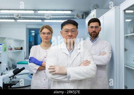 Porträt eines jungen Teams von Wissenschaftlern und Ärzten, die in einem modernen Labor in weißen Mänteln und Schutzausrüstung stehen, ihre Arme kreuzen und ernsthaft die Kamera betrachten. Stockfoto