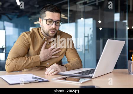 Panic attack in the workplace. A young male office worker sits at a desk and holds his chest, suffers from heavy breathing, sweating. Stock Photo