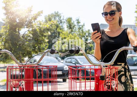 Junge städtische Frau in Freizeitkleidung und Brillen, die Fahrrad mit ihrem Telefon und Stadträder-Sharing-System mietet. Stockfoto