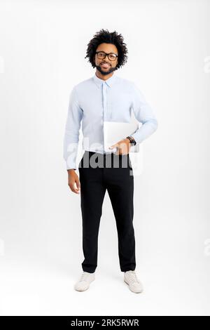 Foto in voller Länge von einem fröhlichen jungen Mann, der einen Laptop auf weißem Hintergrund hält, Geschäftsmann, der mit einem glücklichen Lächeln auf die Kamera schaut. Männlicher Mitarbeiter oder Freiberufler, der einen Laptop mit sich führt. Leerzeichen kopieren Stockfoto