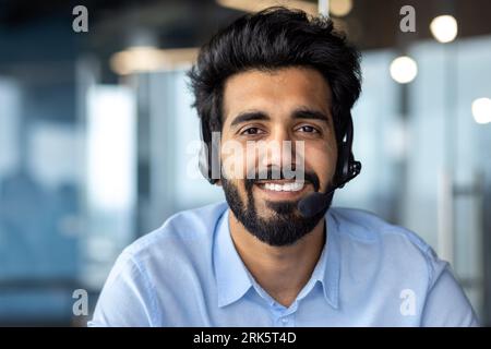 Close-up photo. Portrait of young Indian male student wearing headset sitting in campus office, studying online, smiling at camera. Stockfoto