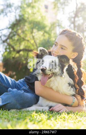 Vertical portrait of a smiling red-haired girl with braids hugging her black and white dog on the grass in a park. Copy space Stock Photo