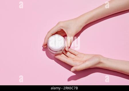 Top view of young tender woman's hands holding open jars of cosmetic creams on pink background. Female face care. Stock Photo