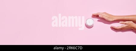 Banner. Top view of young tender woman's hands holding open jars of cosmetic creams on pink background. Female face care. Stock Photo
