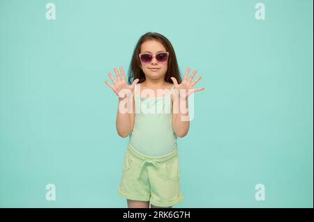 Fröhliches kaukasisches Kind im Grundalter, entzückendes kleines Mädchen im Alter von 6 Jahren, in Sommerkleidung und Sonnenbrille gekleidet, zeigt ihre Hände nach oben, schaut Stockfoto